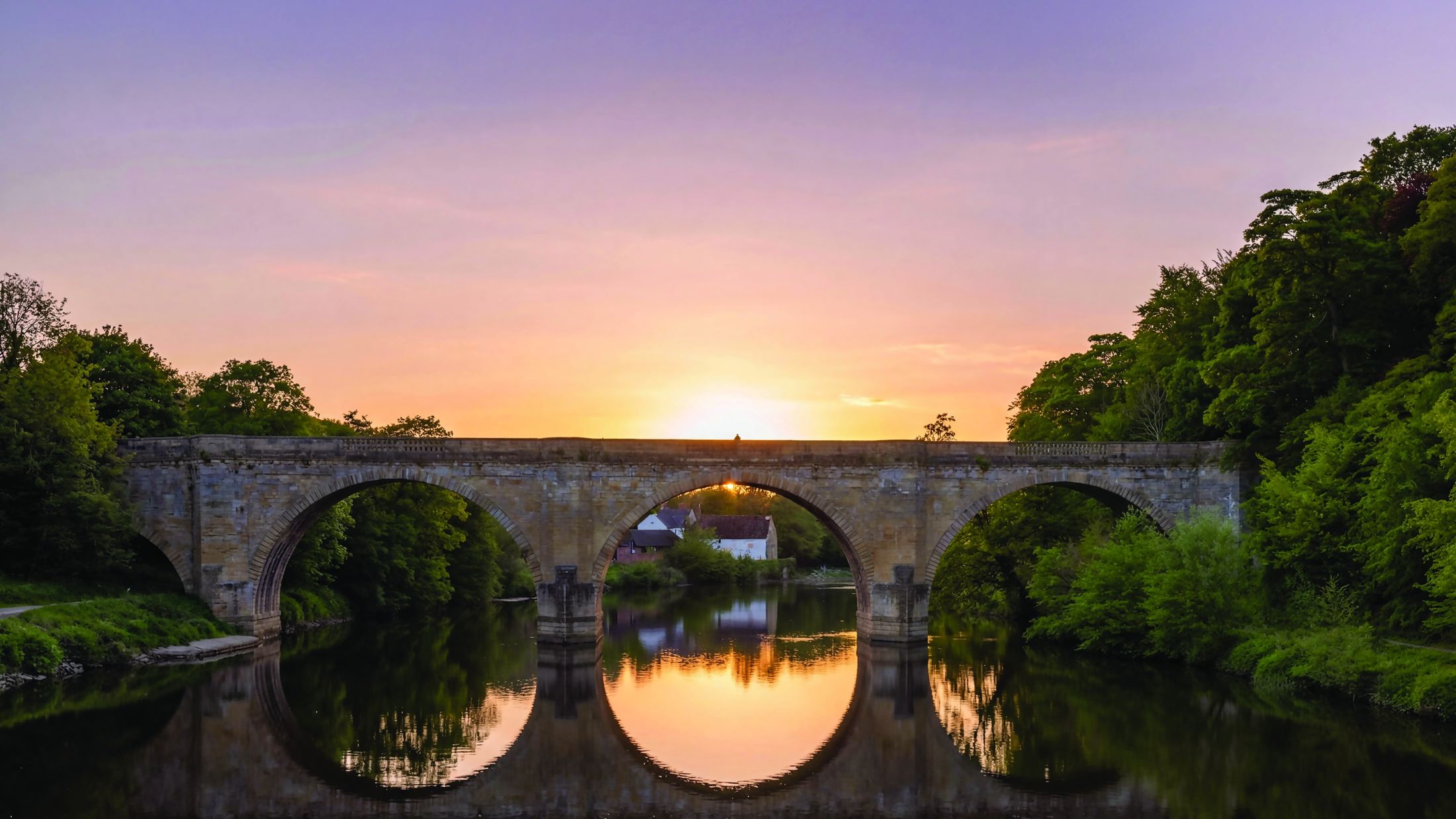 A three-arched stone bridge over a still river at sunset, reflected in the water