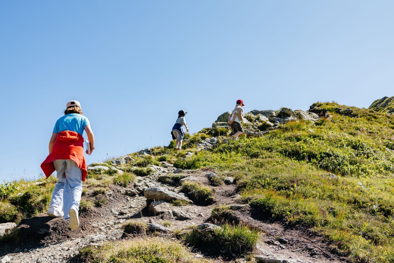 Three children walking up a grassy hillside path under a blue sky